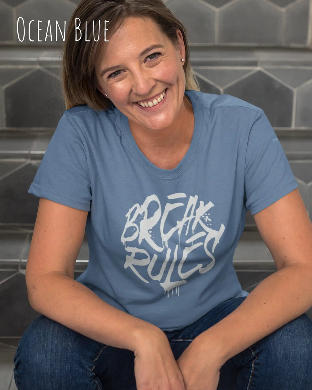 Eine lächelnde frau trägt ein design t-shirt mit femininem schnitt in der farbe ocean blue und mit der grafik "break rules" vorne auf der brust