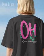 Frau trägt das Heavyweight Oversized T-Shirt AWDis 240 T in der Farbe deep black mit dem Design OH STAY AMAZED auf dem Rücken.