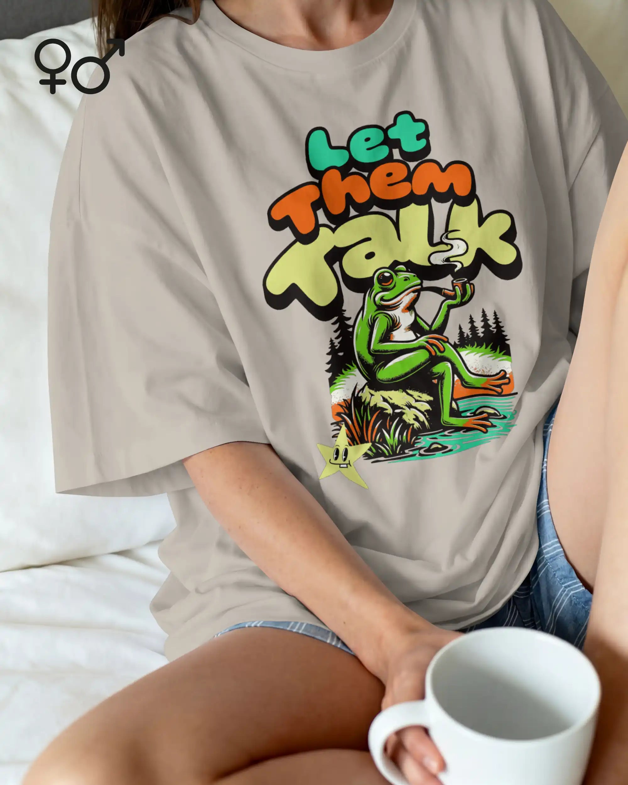 eine frau mit Kaffeetasse trägt ein oversized heavyweight design-t-shirt in der farbe natural stone  mit dem entspannten motiv let them talk - lass sie reden. 