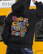 eine frau trägt einen Oversized Design-Hoodie JH111 in  deep black  mit der farbenfrohen grafik "Smile ... it confuses people" auf dem rücken.
